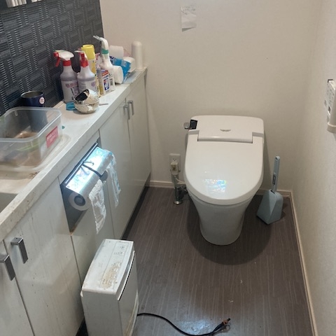 下野市の戸建て住宅トイレにて撮影した清掃前の様子。床にはホコリや物品があり、洗剤や雑品がカウンターに乱雑に置かれている。