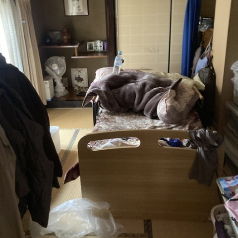 栃木県壬生町の戸建て住宅・片付け前の居室。ベッド周りに衣類や寝具が散乱し、生活導線が遮られている様子。