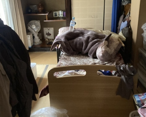 栃木県壬生町の戸建て住宅・片付け前の居室。ベッド周りに衣類や寝具が散乱し、生活導線が遮られている様子。