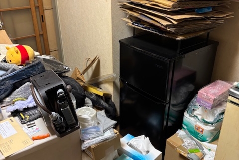 冷蔵庫の上に段ボールや古紙類が積み重なった片付け前の室内の様子。生活用品や雑貨が散在し、整理が必要な状態の1DKアパート