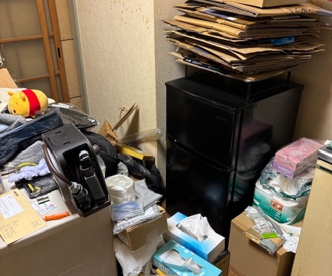 冷蔵庫の上に段ボールや古紙類が積み重なった片付け前の室内の様子。生活用品や雑貨が散在し、整理が必要な状態の1DKアパート
