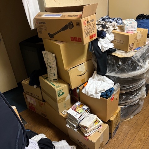 段ボールや衣類、書籍などが山積みになった居室の片付け前の様子。スペースが圧迫され、通路の確保も困難な状態の室内